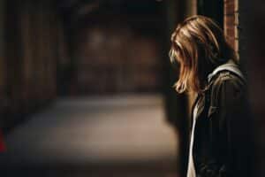 teen girl leaning against wall in dim hallway short blonde hair covering her face and wearing a hoodie